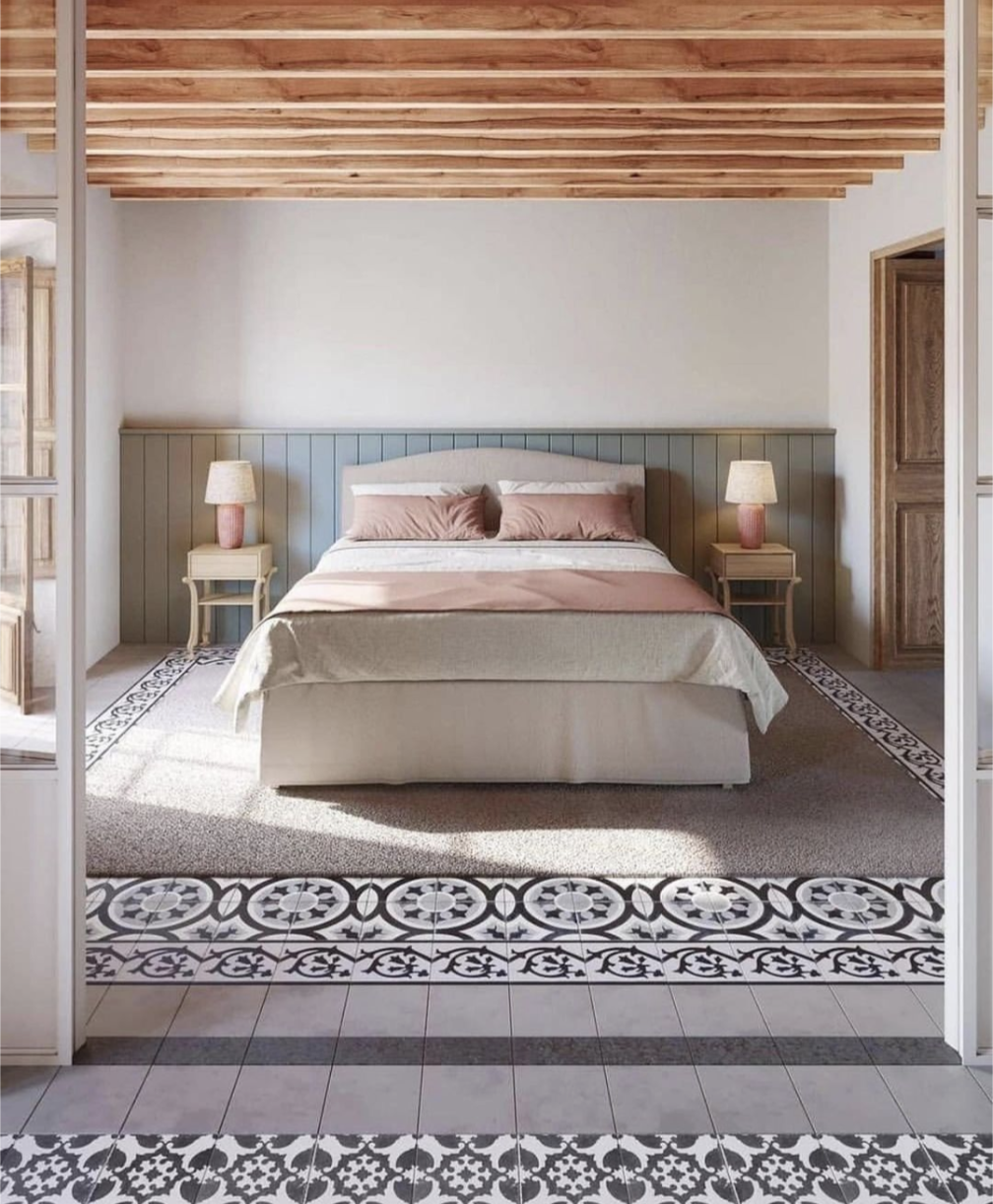 A cozy bedroom with a double bed, wooden ceiling beams, patterned tile floor, and two bedside tables with lamps.
