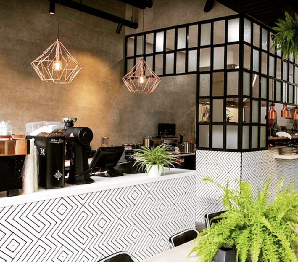 Modern café interior with geometric light fixtures, patterned counter, lush green plants, and a coffee machine on the counter.