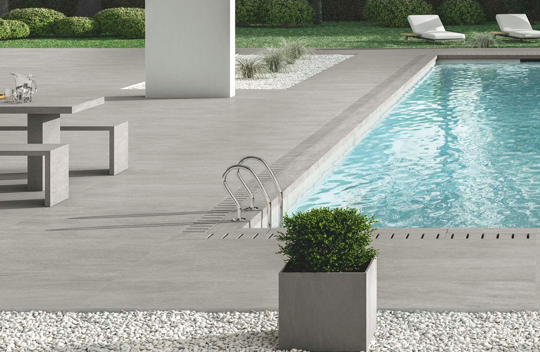 Modern poolside patio with gray tiles, a picnic table, potted plant, and two lounge chairs in a grassy area.