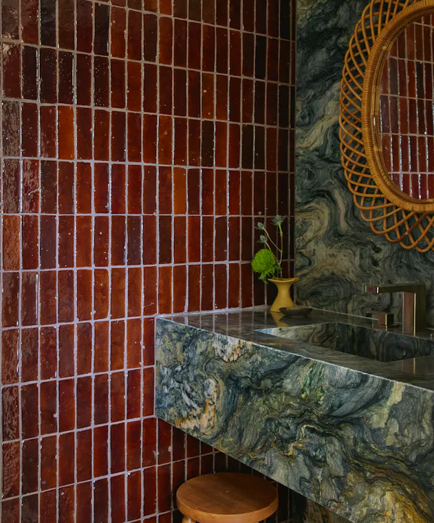 Bathroom with glossy red-brown tiles, a blue-green marble sink, a wooden stool, and a round wicker mirror on the wall.