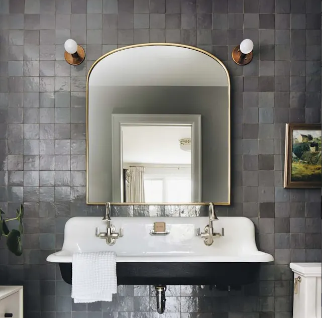 A bathroom with an Ash Grey New York Cement Tile wall, large arched mirror, double faucets, and white sink with towel.