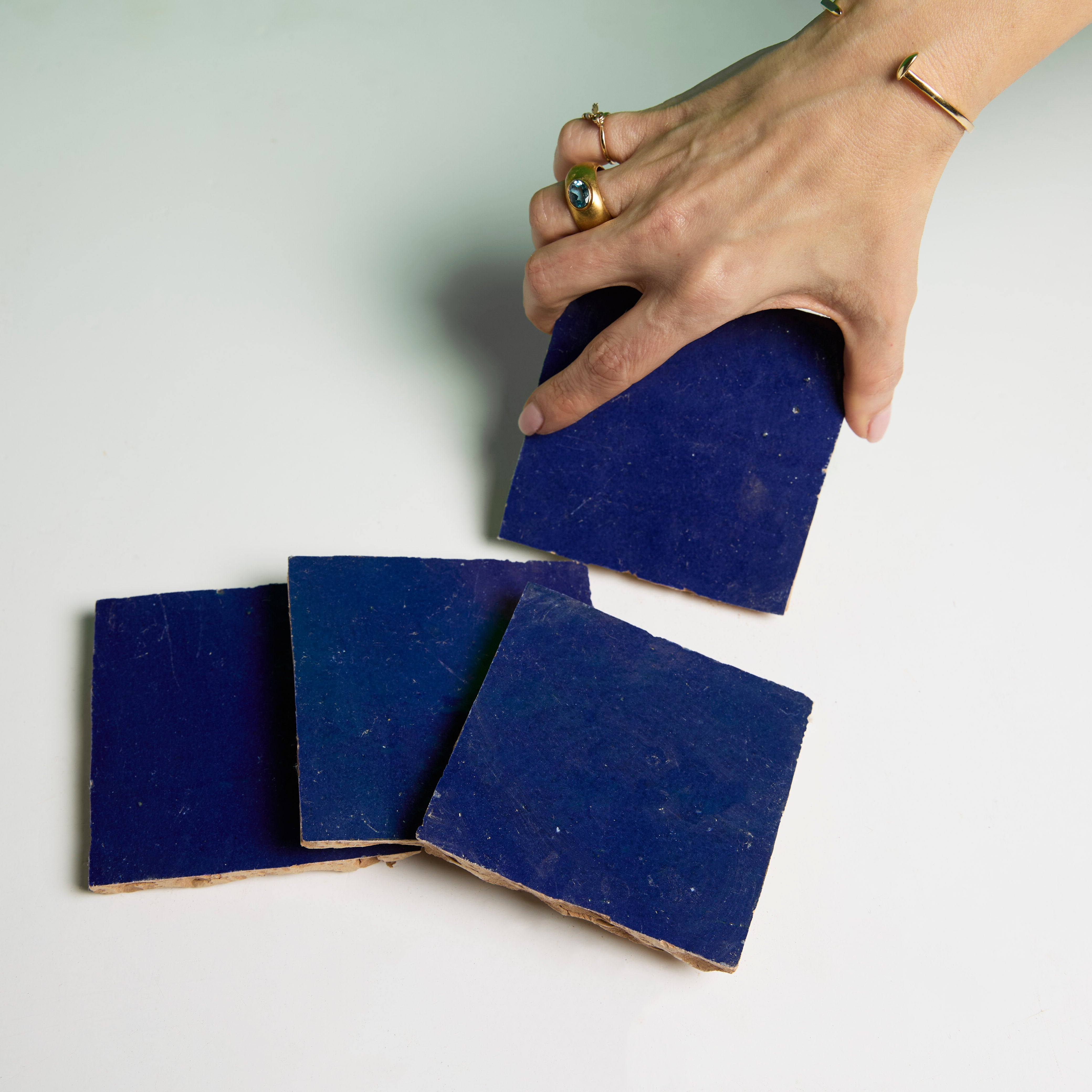 A hand picks up a Navy Depths tile by New York Cement Tile from four dark blue handcrafted pieces on a white surface.