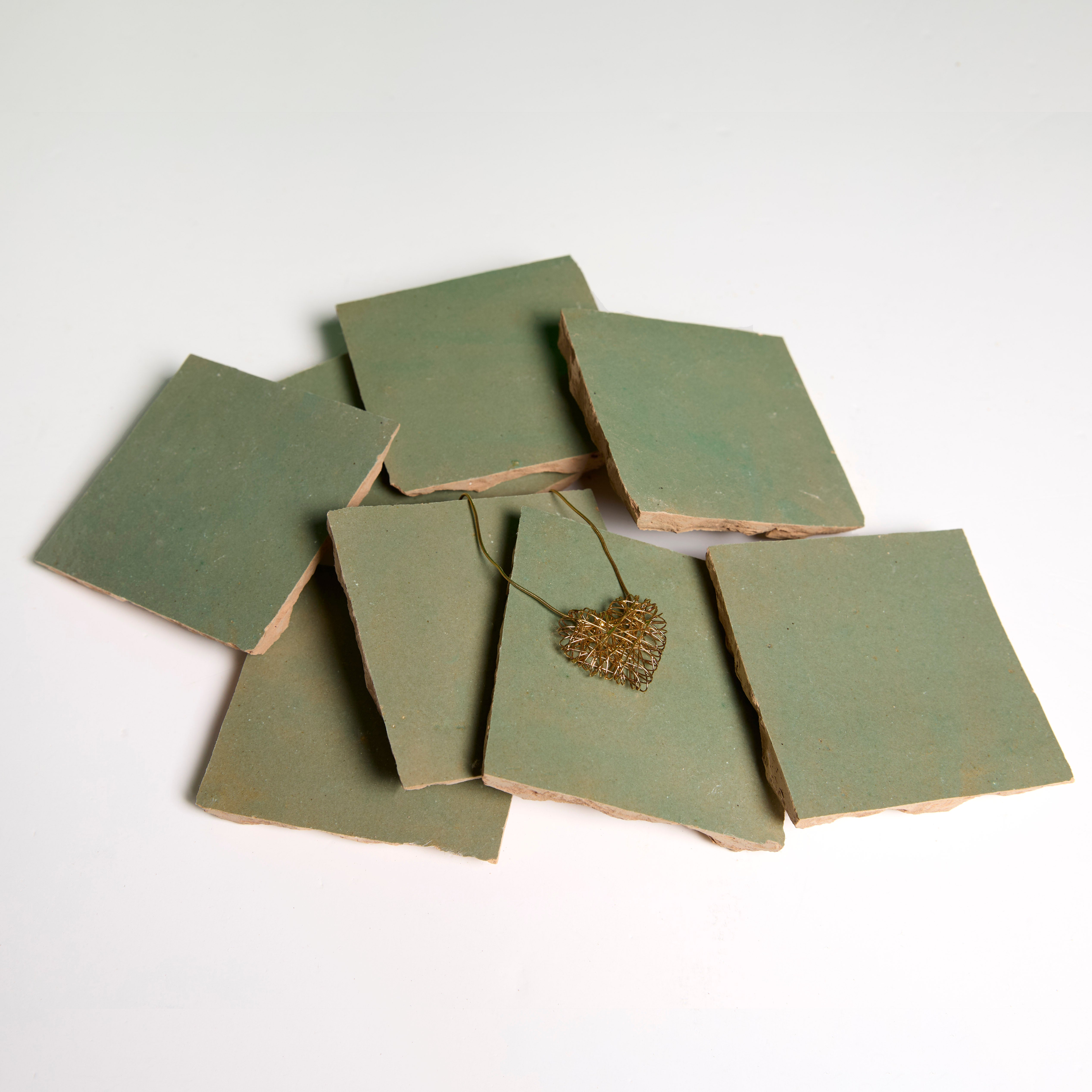 Six Olive Grove tiles by New York Cement Tile, with rough edges, are stacked in a pile and topped with a gold leaf pendant.