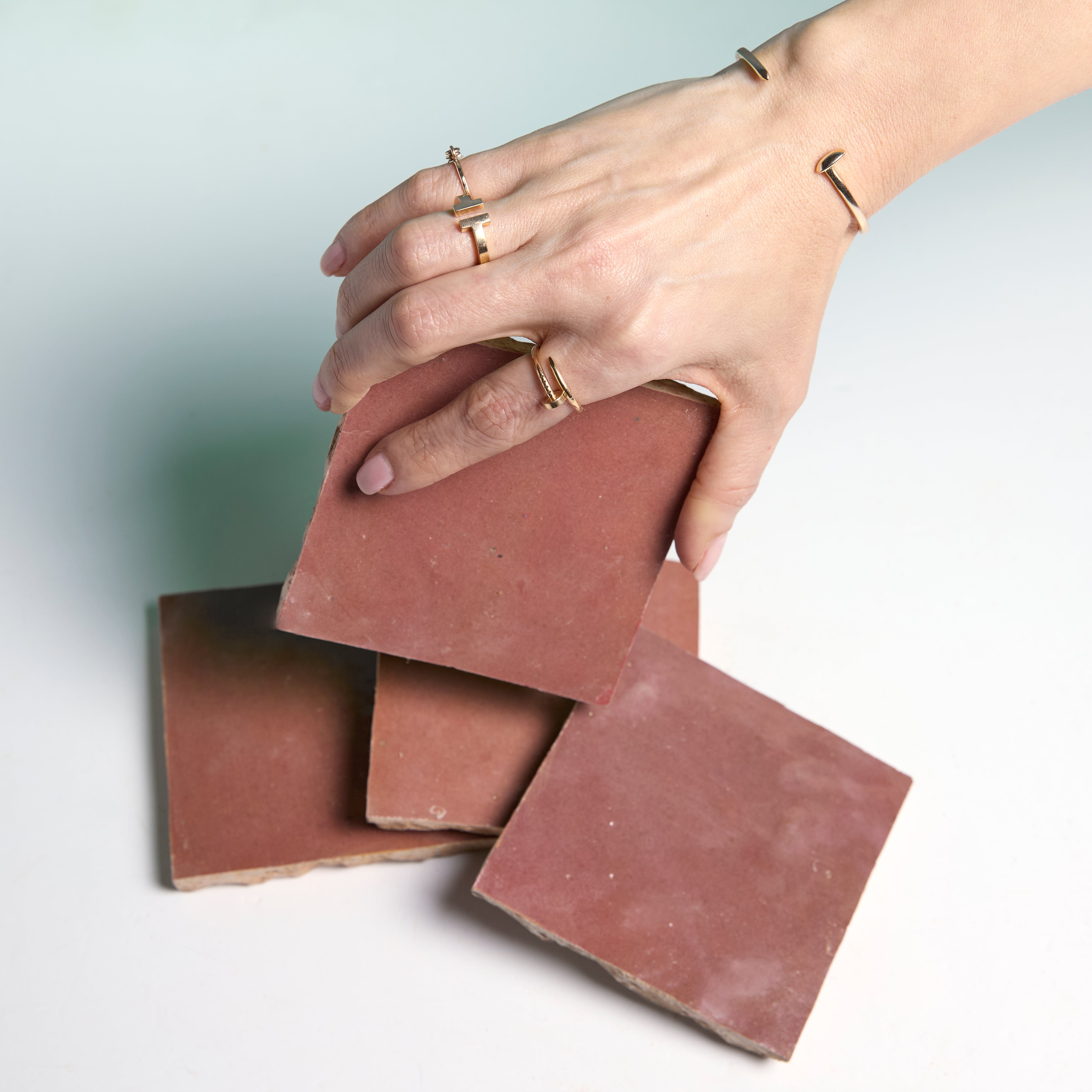 A hand with gold jewelry holds a Glazed Copper Rose tile by New York Cement Tile; three more are stacked below on white.