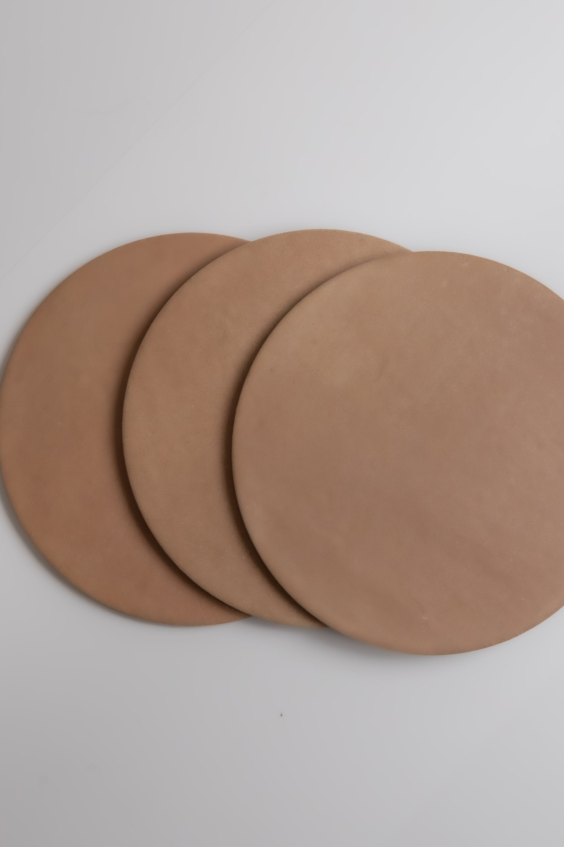 Three round, flat Arcilla samples by Finca, in light brown, overlap on a white surface.