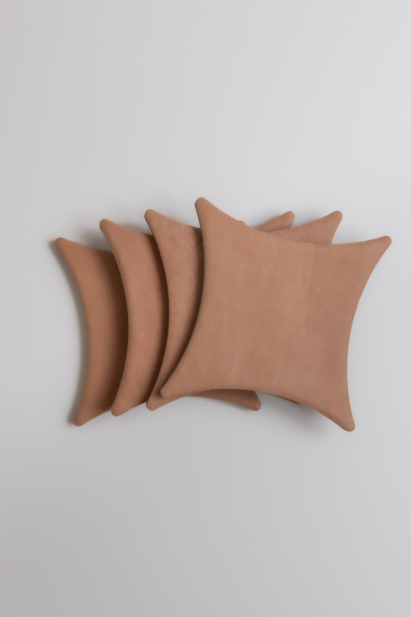 Four tan, square Arcilla Sample cushions by Finca with pointed corners, arranged in a neat overlapping row on white.