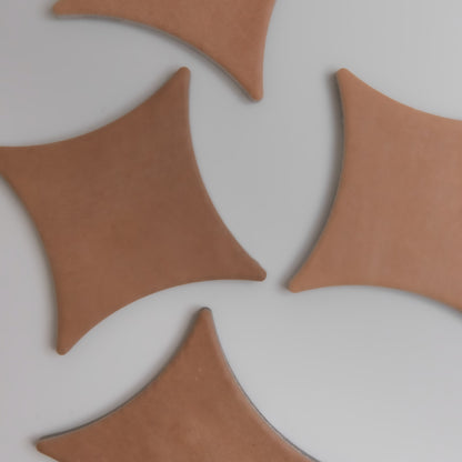 Four brown, curved, diamond-shaped Arcilla tiles by Finca are arranged on a light gray surface with gaps between them.