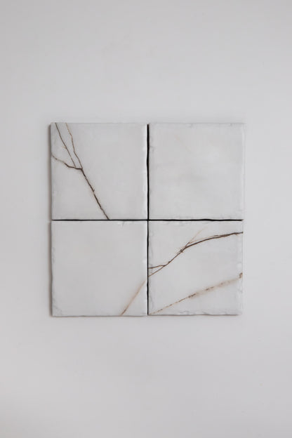 Four Tosca Calacatta Soft sample tiles with light brown veining are arranged in a 2x2 grid on a white wall.