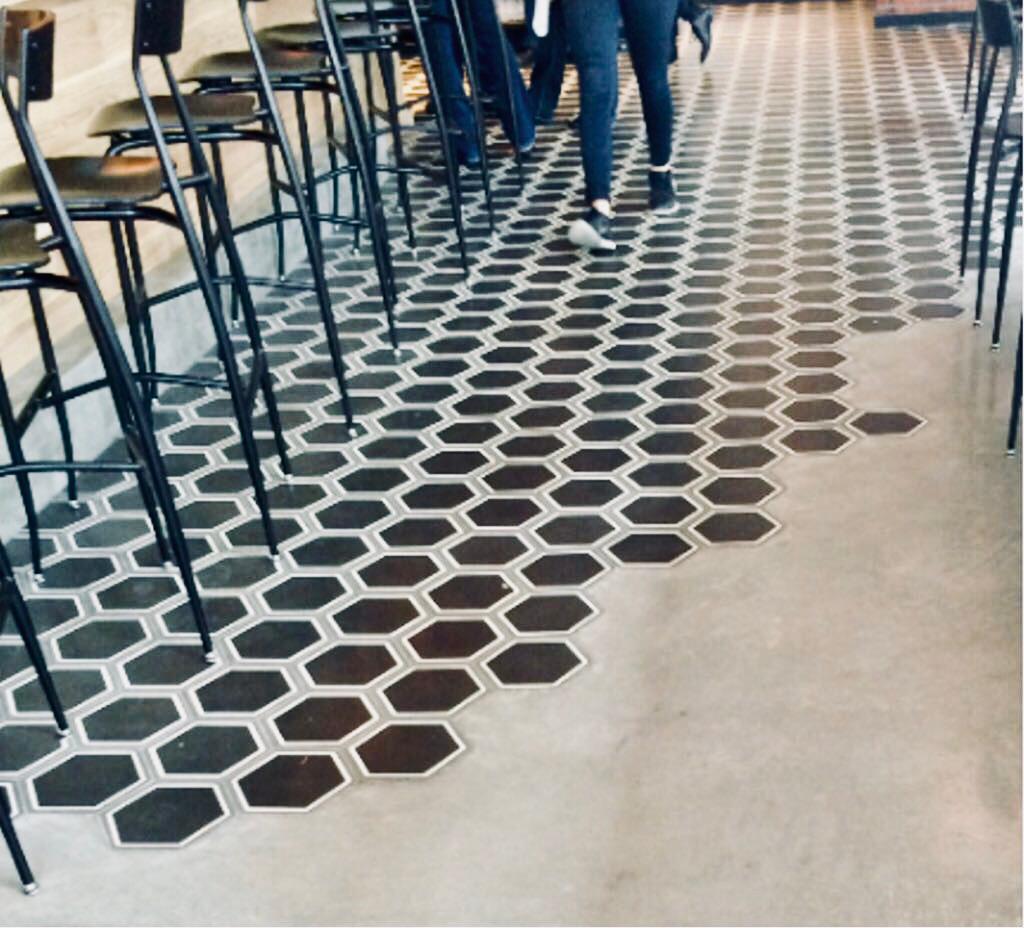 A floor with hexagonal black tiles blending into plain concrete, with people walking and tall chairs along a counter.