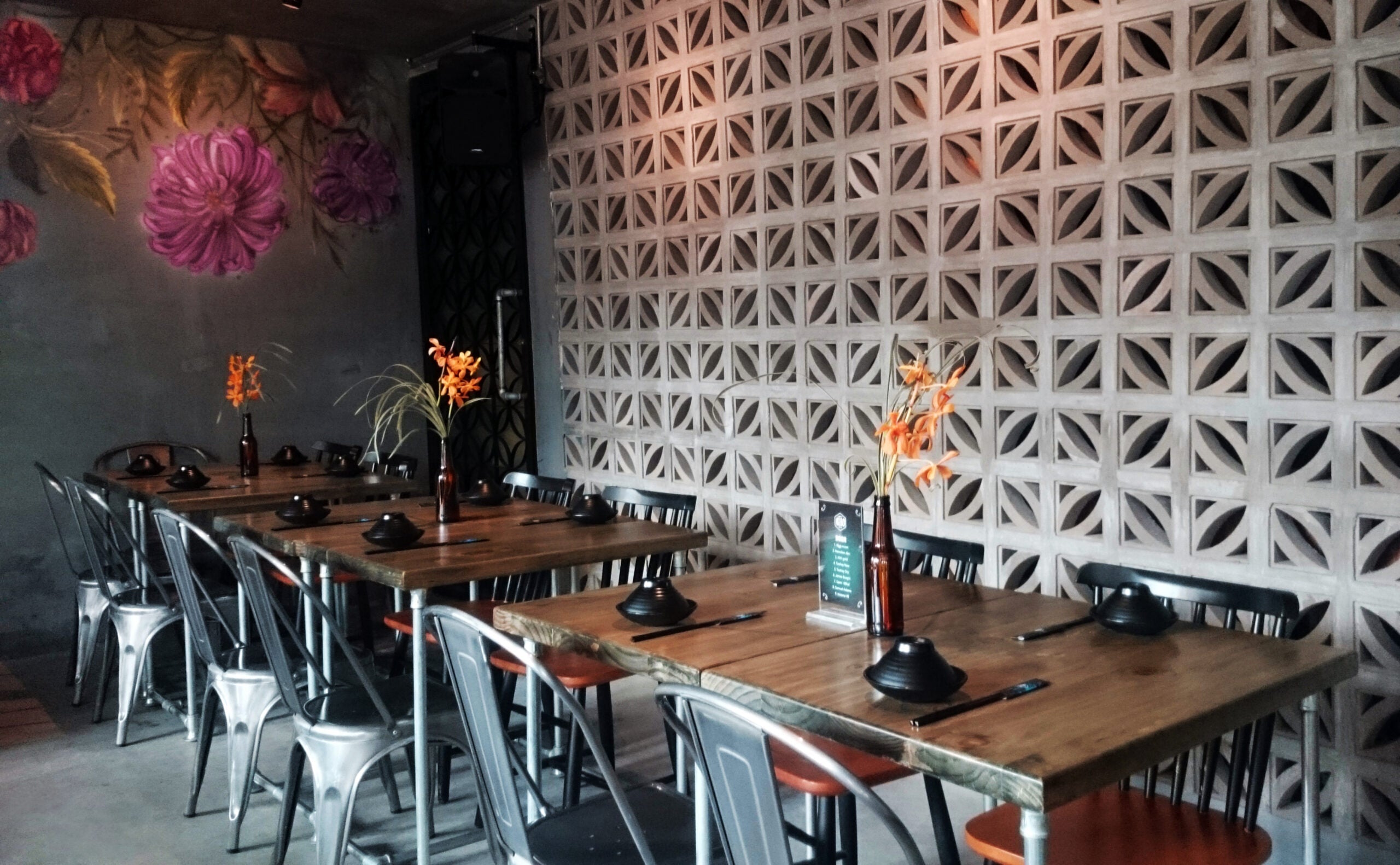 A modern restaurant with wooden tables, metal chairs, floral wall art, and orange flowers in vases on each table.