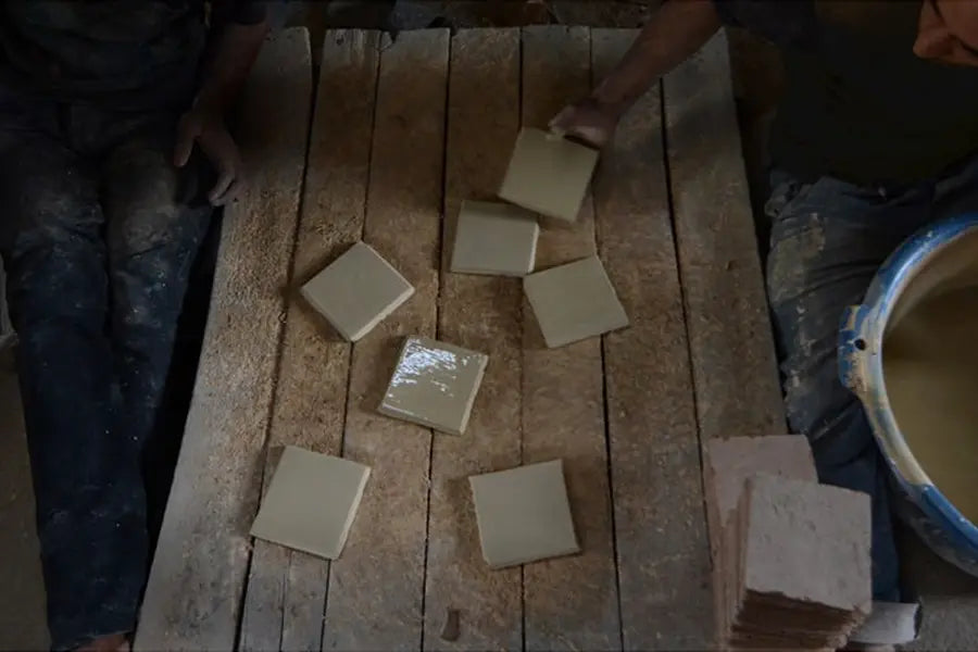 Two people place wet square tiles on a wooden surface beside a stack of tiles and a container of liquid clay.