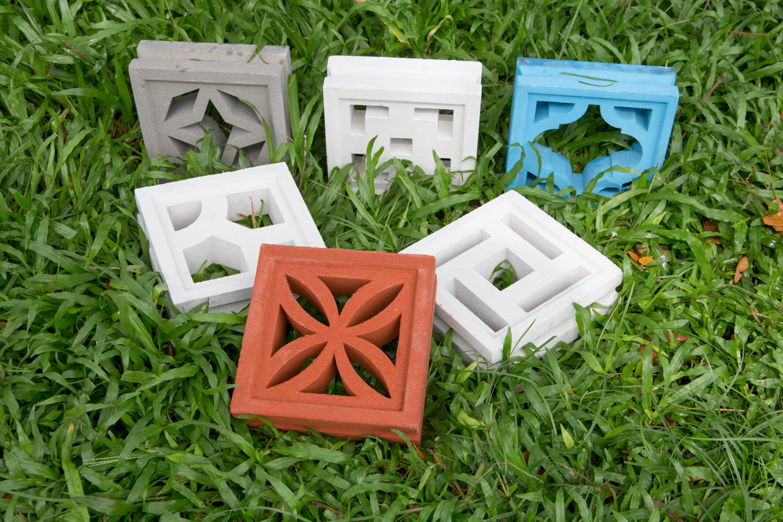 Six Beeza Sample breeze blocks by New York Cement Tile with unique patterns sit on grass; one is red, another blue.