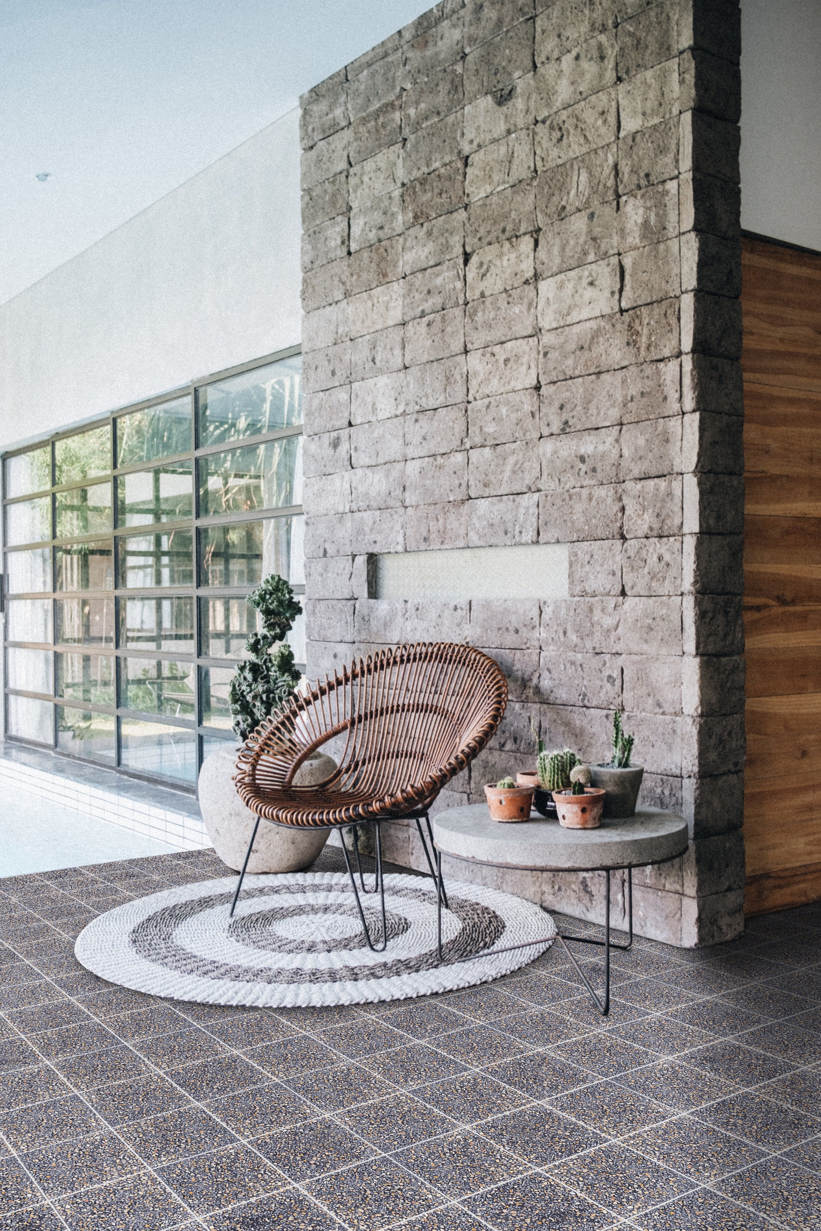 Wicker chair and small table with potted plants on a round rug by a stone wall in a modern, sunlit room.