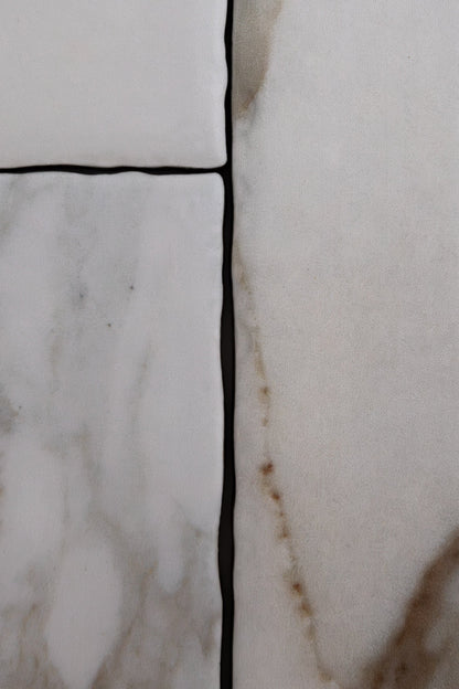 Close-up of Tosca Macchia Vecchia Extra Sample—four white tiles with dark grout and light brown veining on the right.