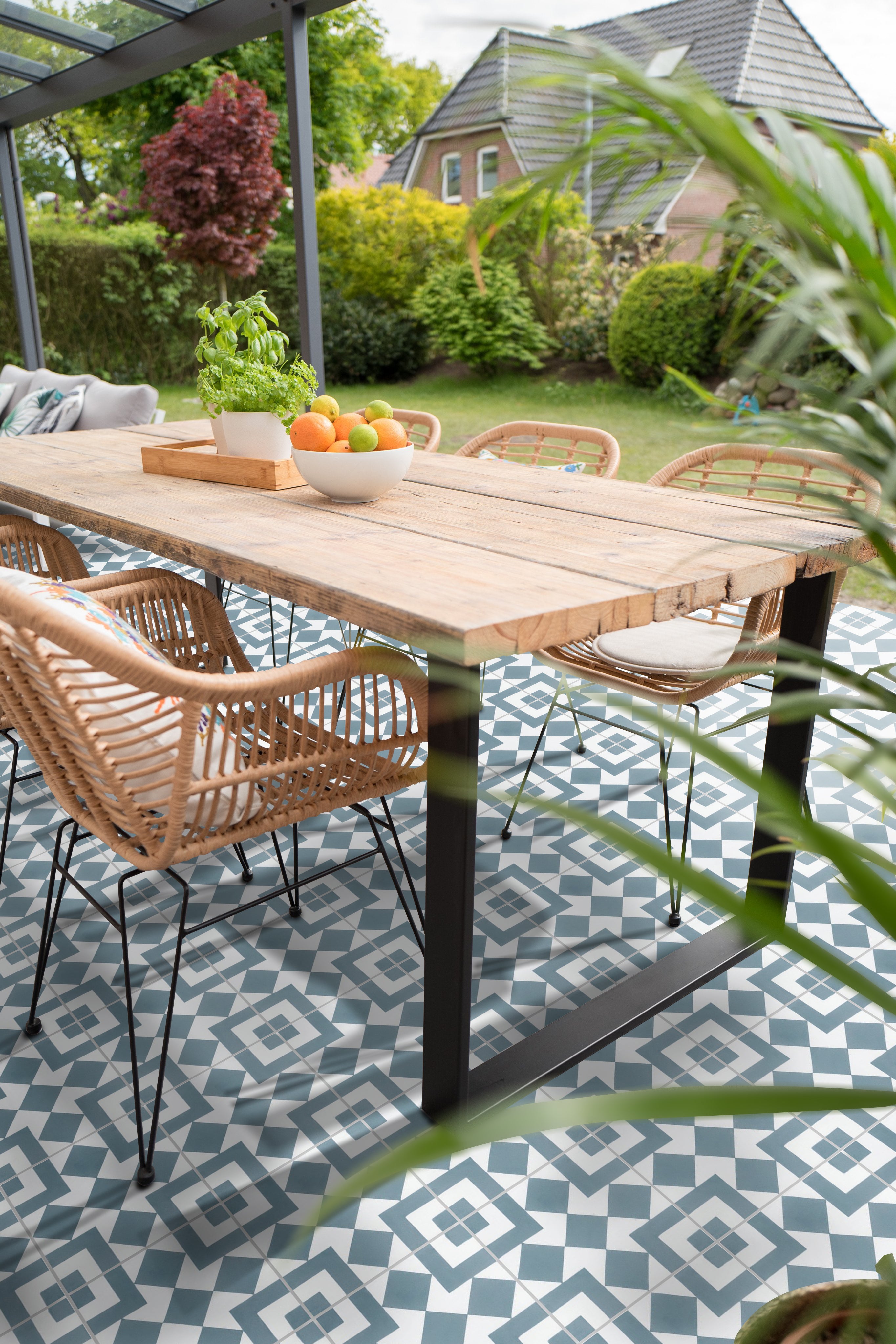 Outdoor dining area with a wooden table, rattan chairs, and geometric patterned tiles, overlooking a green garden.