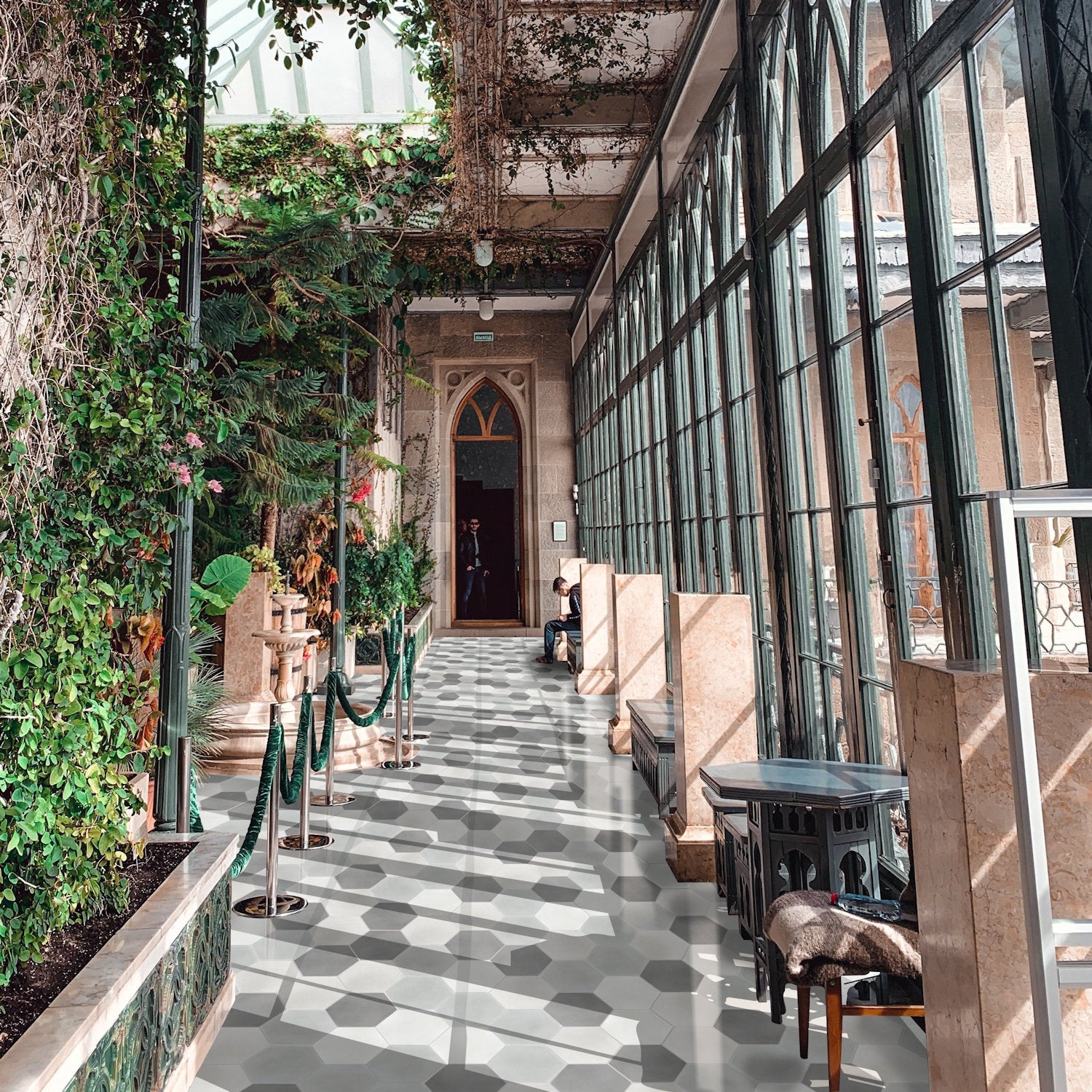 A sunlit glass corridor with green plants, tall windows, and elegant seating on Solid Hex - Mixed Grey by New York Cement Tile.