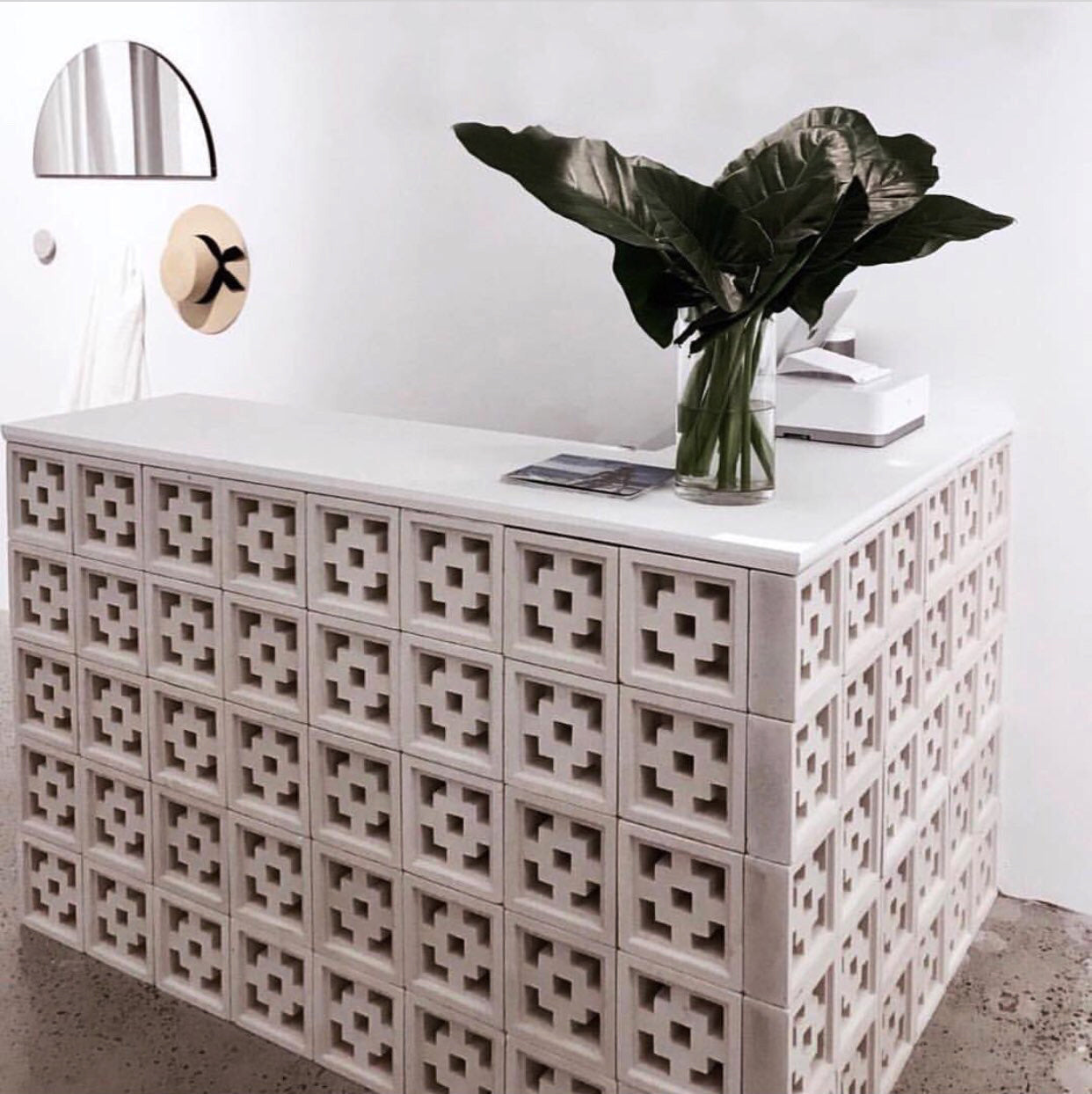 A Tong counter by New York Cement Tile features a vase of green leaves, magazines, and a wall mirror with a hat behind.