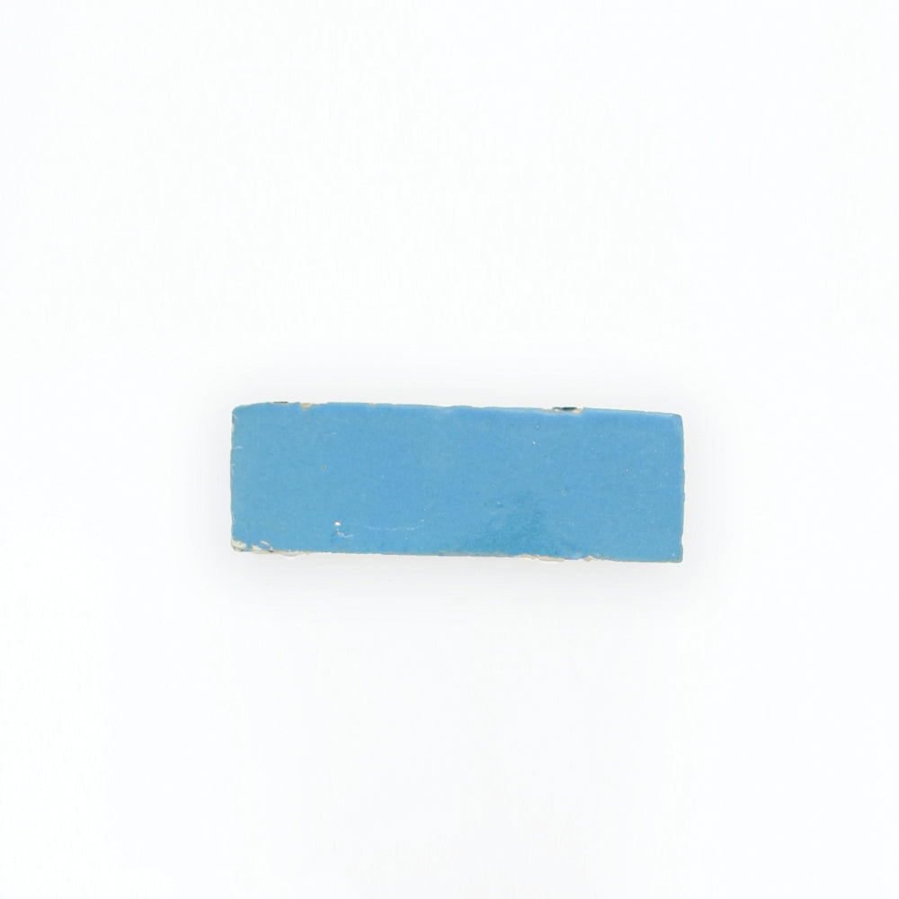 An Azure (2x6) rectangular blue tile by New York Cement Tile rests on a plain white background, edges slightly worn.