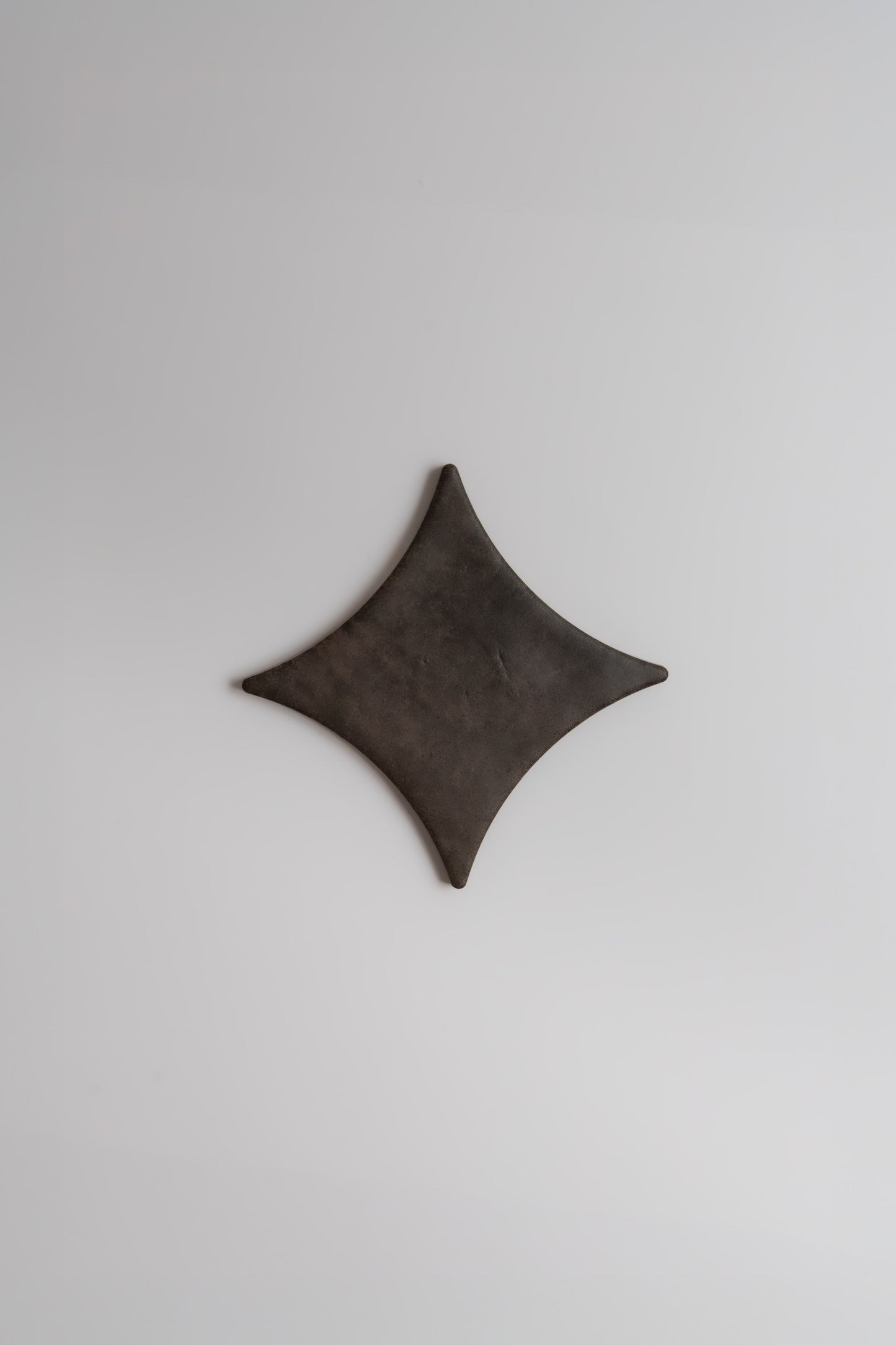 The Finca Ocre, Extra Sample features a dark, diamond-shaped form with curved edges on a plain light gray background.