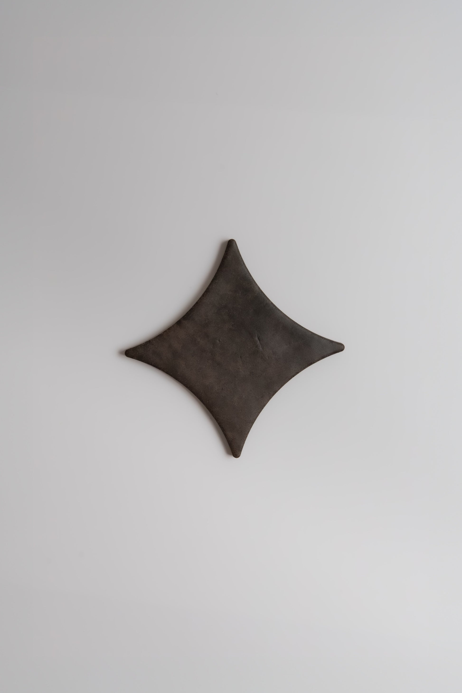 The Finca Ocre, Extra Sample features a dark, diamond-shaped form with curved edges on a plain light gray background.