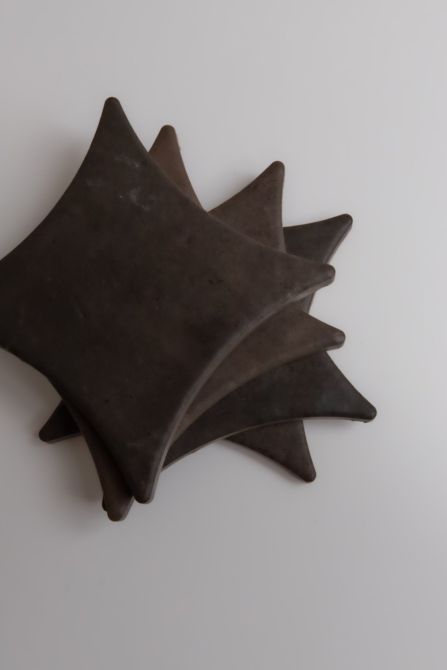 Four dark, square Ocre, Extra Sample coasters by Finca stack in a spiral on a light surface.