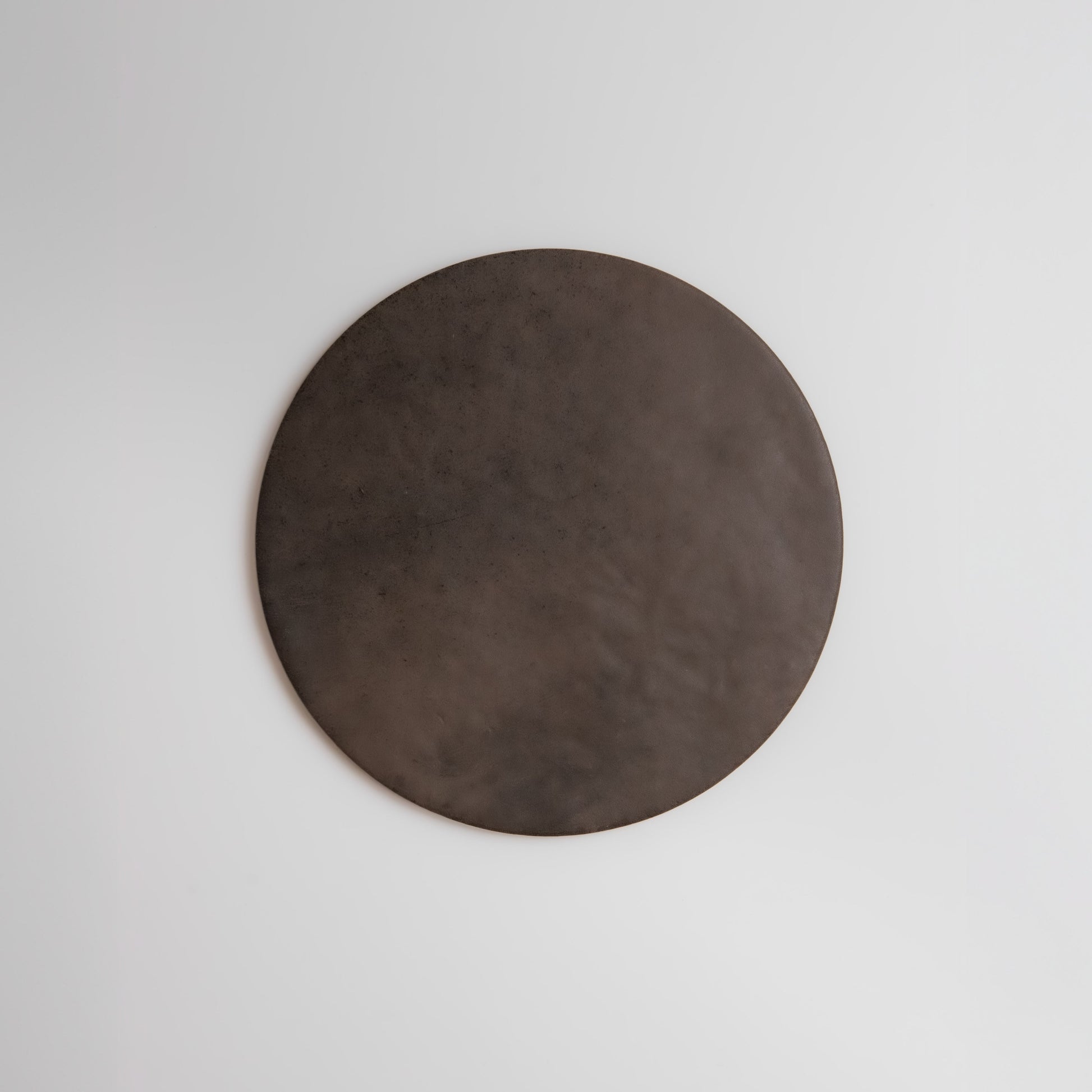 The Ocre, Extra Sample by Finca is a large, flat, dark brown circle displayed on a plain white wall.