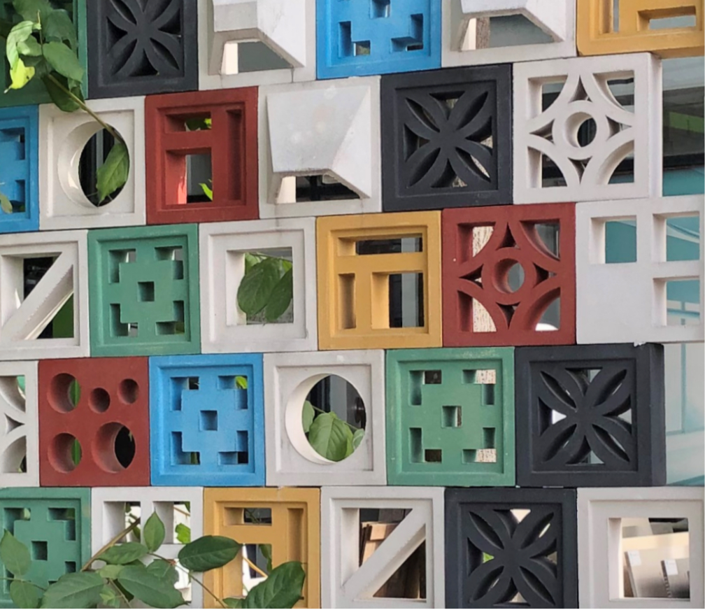 Colorful decorative concrete blocks with geometric patterns form a wall, with green leaves peeking through some openings.