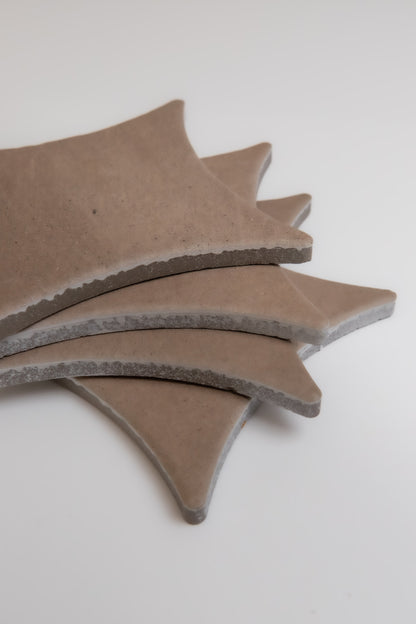 Four tan Pampas tiles by Finca, with curved shapes and rough edges, are stacked on a white surface.