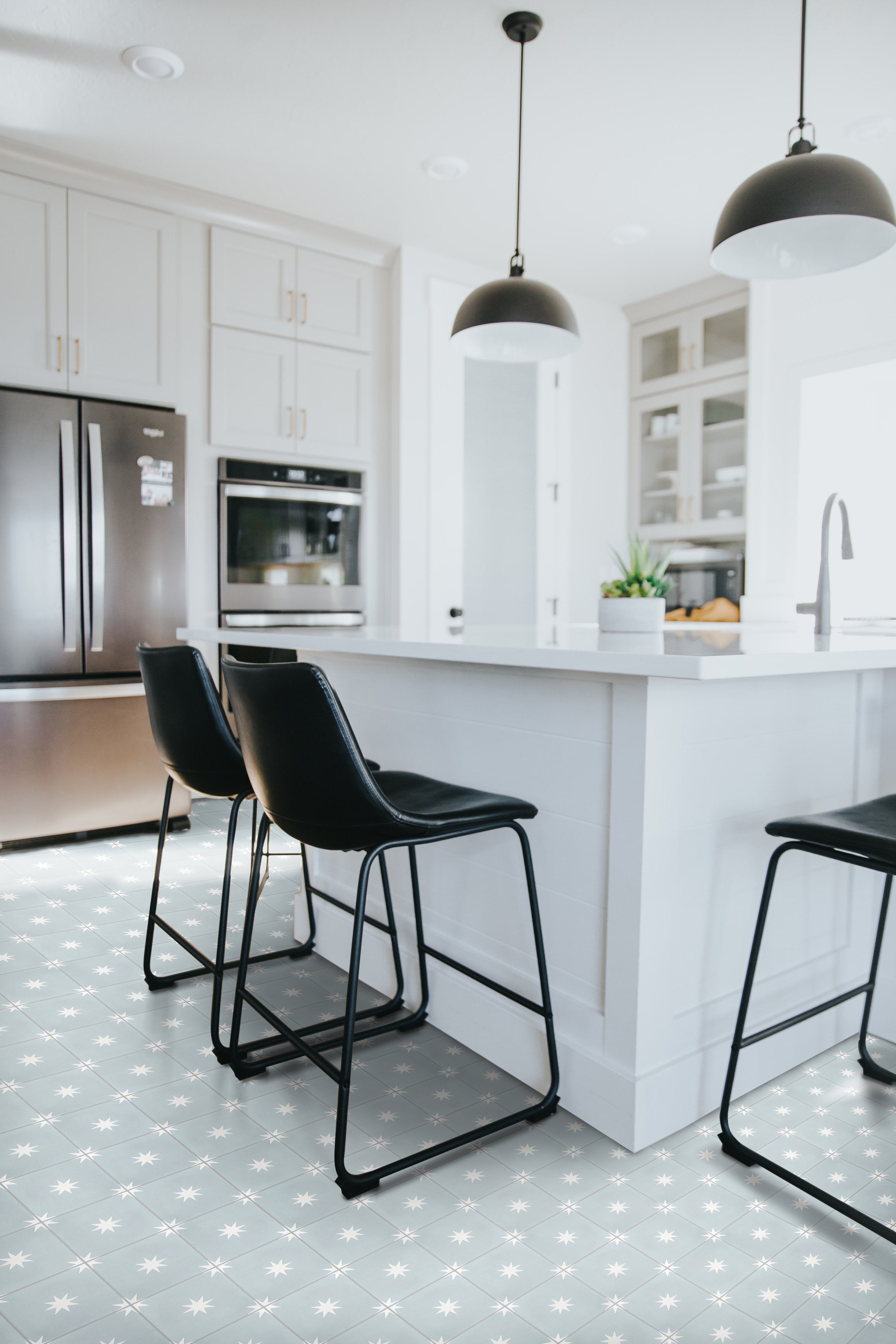 Modern kitchen with black bar stools, white island, pendant lights, and stainless steel appliances on patterned tile floor.