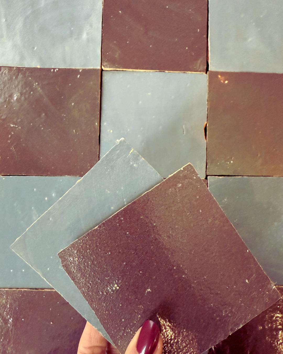 A hand holds two Thistle tiles by New York Cement Tile over a checkered brown and blue-grey tile floor.